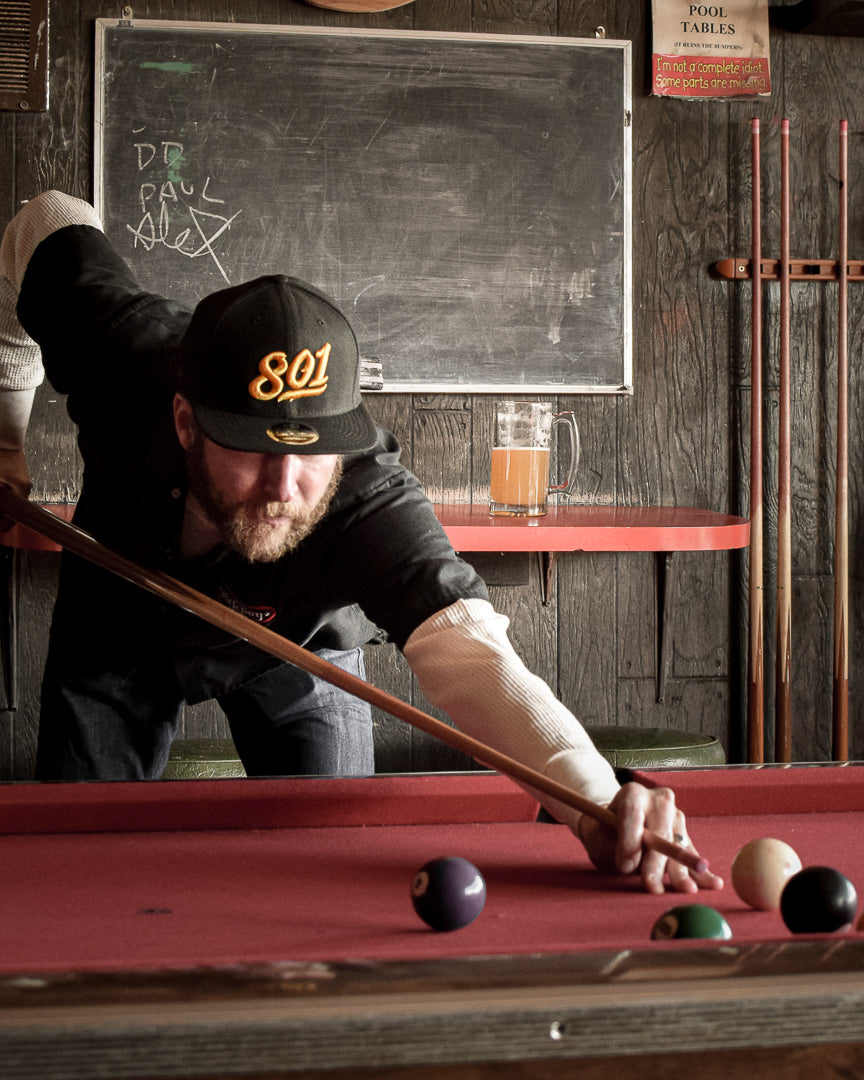 A man in a Uinta 801 hat shoots pool in a barroom