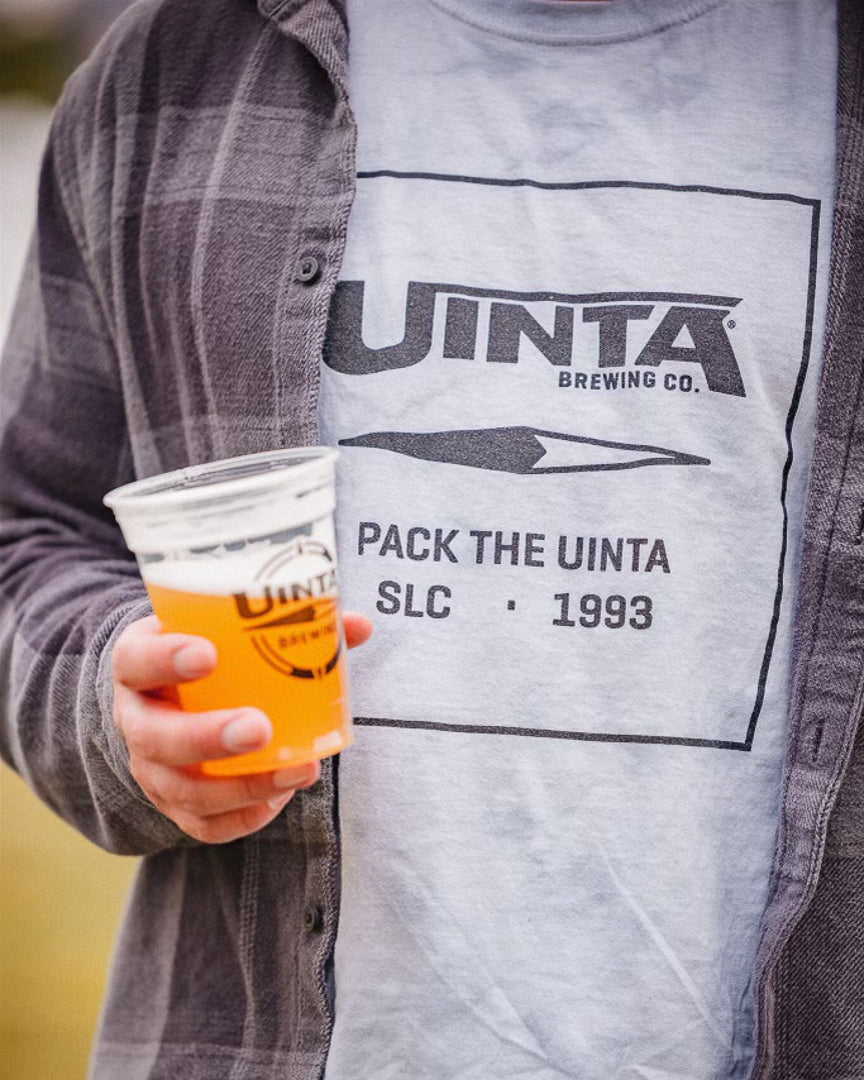 A cropped view of a person holding a plastic cup of beer, wearing a flannel shirt and Uinta tee
