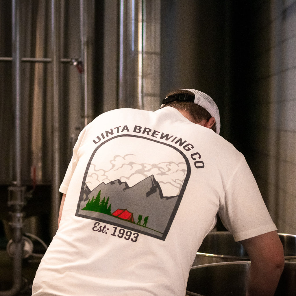 A brewer wearing a t-shirt with a mountain graphic reaches into a brewing kettle