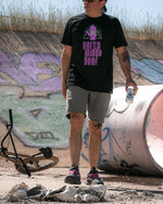 Load image into Gallery viewer, A person  holds a can of beer while walking near a bicycle and a graffiti covered backdrop
