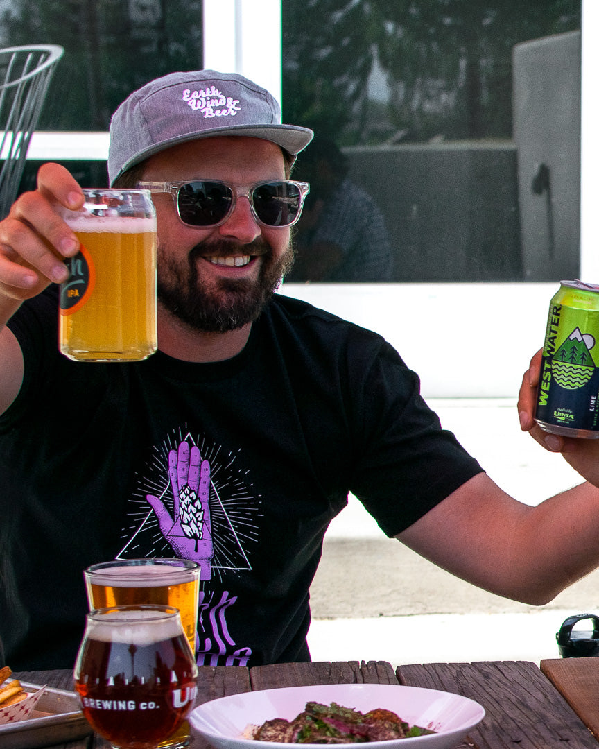 A smiling man wearing a hat and sunglasses raises a glass of beer 